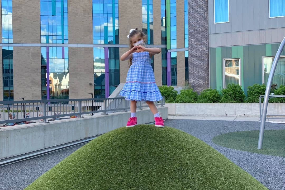 Young girl in blue dress making a heart with her hands on a grass hill.