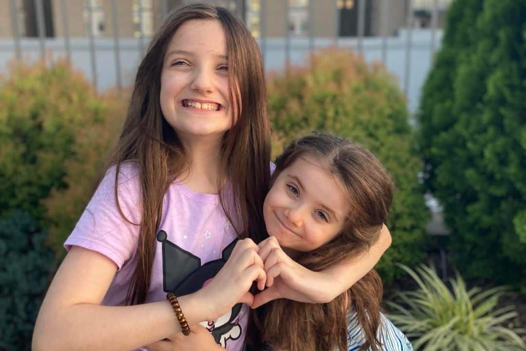 Big sister hugging little sister and making a heart with her hands outside.